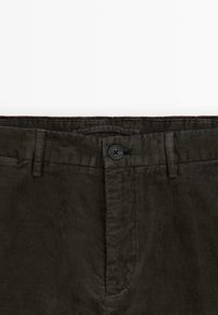 Pantalon en velours côtelé vert foncé avec passants de ceinture, fermeture par un seul bouton et braguette zippée à l’avant à la taille.
