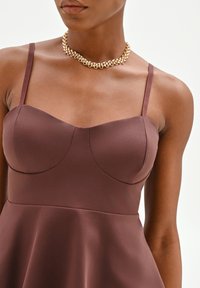 Mujer con un vestido marrón sin mangas con tirantes finos y un collar grueso de cadena dorada sobre un fondo liso.