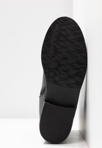 Semelle d'une chaussure en cuir noir avec motif de grip texturé, taille 37 marquée sur la zone de la voûte plantaire, photographiée en position verticale sur un fond blanc.