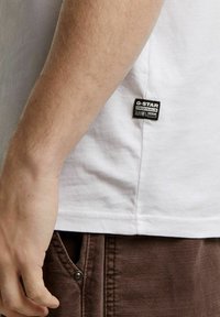 T-shirt bianco in cotone con una piccola etichetta nera G-Star sul lato, abbinato a pantaloni marroni con tasca a chiusura a scatto.