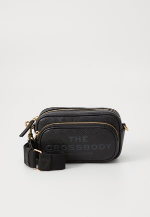 Čierna kožená crossbody taška so zlatými zipsami, nastaviteľným popruhom a embosovaným textom "THE CROSSBODY" na prednej strane.