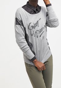 Pull gris avec un texte en sequins, des accents en dentelle sur les côtés et des poignets côtelés, associé à une chemise noire à col et un pantalon vert olive.