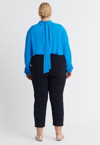 Blaue Bluse mit einem zurückgebundenen Detail, kombiniert mit schwarzen, figurbetonten Hosen mit umgeschlagenen Säumen. Glatter Stoff mit einer eleganten Silhouette.