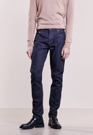 Jeans de mezclilla oscura con un ajuste slim, costuras en naranja contrastante, diseño de cinco bolsillos, y combinados con botas de cuero negras.