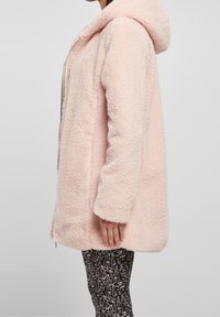 Abrigo de piel sintética rosa con capucha, con cierre de cremallera en la parte delantera y mangas largas. La tela tiene una textura suave y esponjosa. Usado sobre leggings negros con estampado.