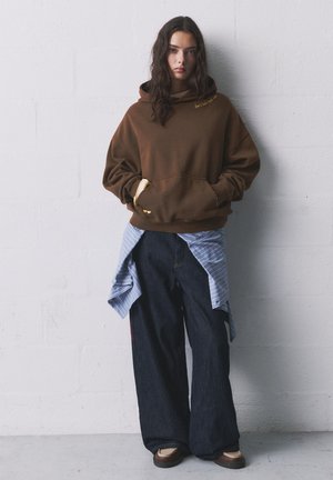 Hanorac maro supradimensionat cu accente galbene, asortat cu blugi negri cu picioare late și o cămașă cu dungi albastre legată în jurul taliei.