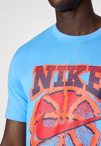 Lys blå bomulds t-shirt med stort, iøjnefaldende orange og sort Nike-logo samt en basketball grafik. Rund hals og korte ærmer.
