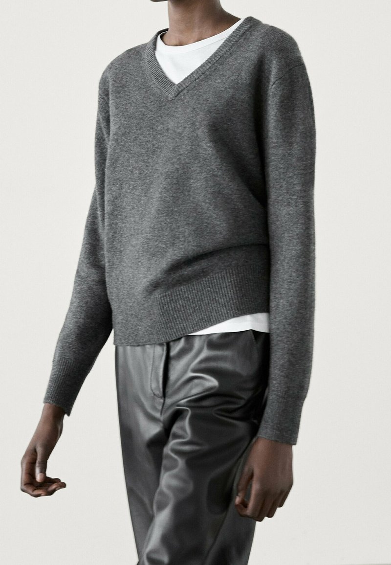 Personne portant un pull gris à col en V superposé sur une chemise blanche, associé à un pantalon en cuir noir, sur un fond clair uni.