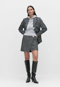 Grå herringbone-blazer över en grå zip-hoodie, parat med en grå omslagsskjol och svarta knähöga stövlar. Släta texturer och strukturerad design.