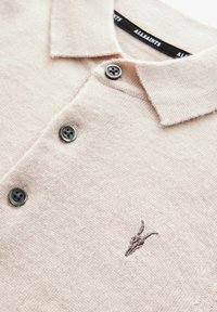 Bledě růžové pletené polo tričko s límečkem, které má tři tmavozelené knoflíky a hnědé logo z rohu vyšívané na hrudi.