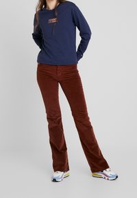 Námořnická dlouhá rukávová košile s logem "TOMMY JEANS", spárovaná s hnědými manšestrovými kalhotami s rozšířenými nohavicemi a barevnými sportovními botami. Hladká textura po celém povrchu.