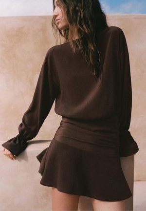 Kvinde iført en mørkebrun ribbet bluse med lange ærmer og matchende høj-taljede nederdel med flæser, lænende sig mod en lys stensurface.