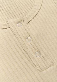 Ribbad beige topp med rund halsringning, tre silverfärgade knäppningar och en liten fickdetalj på framsidan.