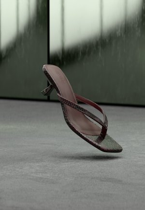 Sandale à talon marron à texture peau de serpent avec fines lanières, suspendue au-dessus d’un sol moquetté gris sur un fond sombre et flou.