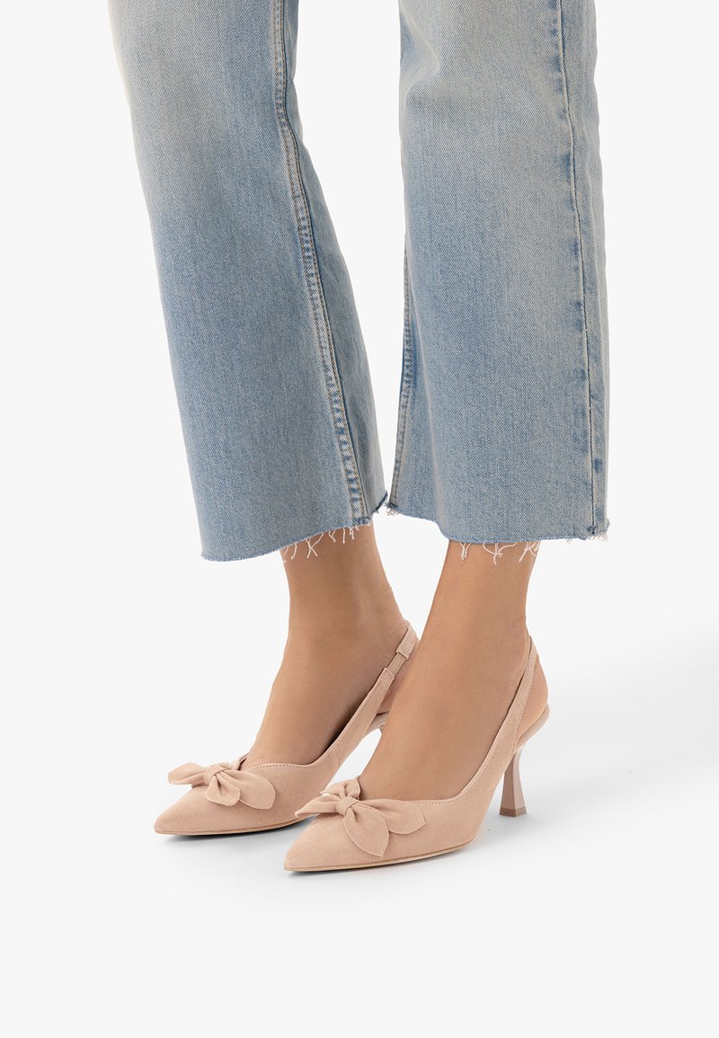 Pieds chaussés de hauts talons en daim beige clair avec des détails en nœud, associés à un jean bleu clair court et effiloché.