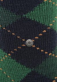 Calcetín de punto con un patrón de argyle verde y azul marino con acentos naranjas, y un emblema de marca metálico en el centro.