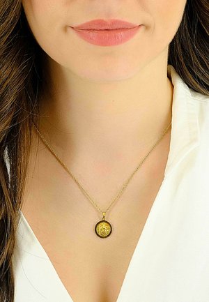 Collier pendentif en or avec un charme circulaire détaillé, présentant un design en relief, suspendu à une chaîne délicate. Porté sur un chemisier blanc.