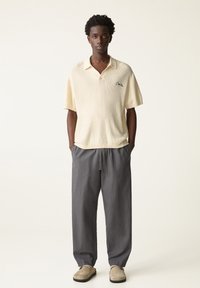 Hellbeige Poloshirt mit kurzen Ärmeln, strukturiertem Strick und gesticktem Detail, kombiniert mit grauen Jogginghosen mit Kordelzug und beigen Slippern.