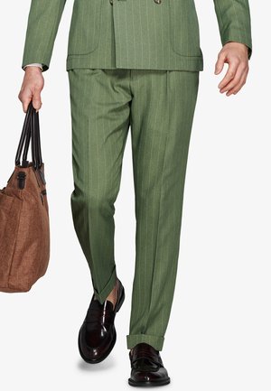 Uomo con pantaloni e giacca a righe verdi, che tiene una borsa a tracolla marrone, camminando con mocassini in pelle marrone scuro.