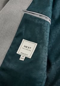 Étiquette sur la doublure sombre à motifs du costume indiquant « NEXT tailoring, regular fit, 40R, made in Bangladesh » à côté d’un tissu gris clair texturé.