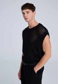 Top senza maniche lavorato a maglia nera con un design morbido e un motivo a rete aperta. Il modello indossa una cintura nera e pantaloni neri su misura.
