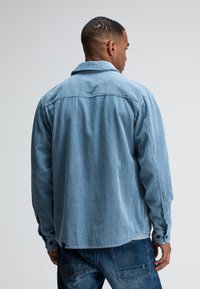 Light blue corduroy jacket with vertical stripes, button cuffs, and a relaxed fit. Features a back yoke and contrasting fabric textures.