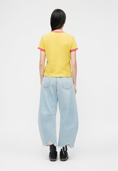 Γυναίκα με μακριά σκουρόχρωμα μαλλιά, φορώντας κίτρινη μπλούζα με ροζ λεπτομέρειες, φαρδιά ανοιχτό μπλε κοντυμένα τζιν και μαύρα αθλητικά παπούτσια, όρθια με την πλάτη στραμμένη.