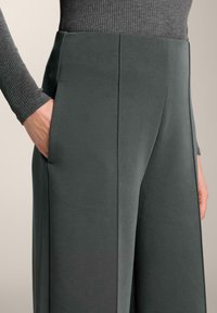 Breda byxor i en mörkgrön tyg, med sidofickor och en smidig textur. Designen har rena linjer och minimal detaljering.