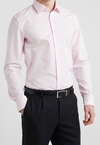 Camisa de manga larga en rosa claro con botones, cuello estructurado y un detalle blanco en el interior del cuello, combinada con pantalones negros.