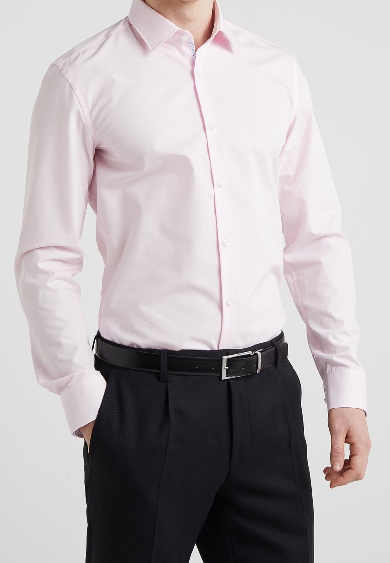 Camisa de manga larga en rosa claro con botones, cuello estructurado y un detalle blanco en el interior del cuello, combinada con pantalones negros.