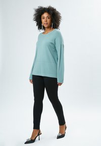 Maglione oversize verde menta con maniche lunghe, abbinato a leggings neri aderenti e décolleté neri con accenti decorativi.