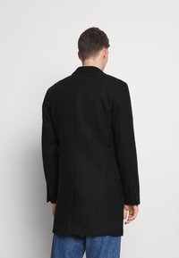 Černý vzorovaný kabát s dlouhým střihem, klopovým límcem s výstřihem do tvaru V a knoflíkovými akcenty na rukávech; texturovaná látka; nošený přes modré džíny.