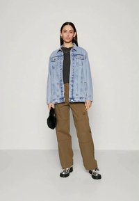 Young woman stands wearing light blue denim jacket, black shirt, brown cargo pants, silver platform shoes, and holding small black bag.