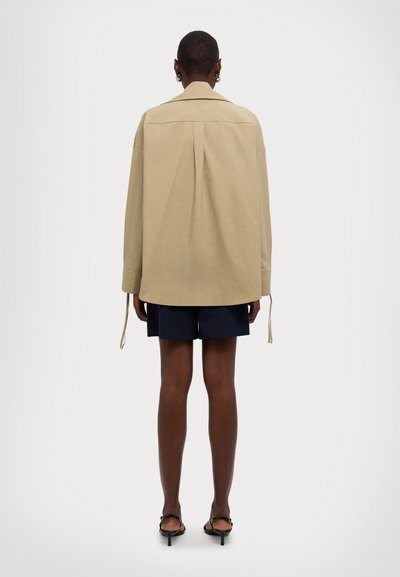 Giacca over-size beige con collo a pieghe, maniche lunghe e legature regolabili. Abbinata a pantaloni corti blu navy e sandali neri minimalisti.
