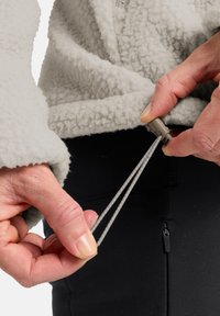Veste en polaire gris clair, avec un tissu texturé. Mains ajustant les cordons élastiques à la taille, associée à un pantalon noir.