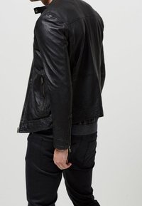 Svart läderjacka med dragkedjestängning, sidofickor och en lätt texturerad yta, bärs över mörka byxor.