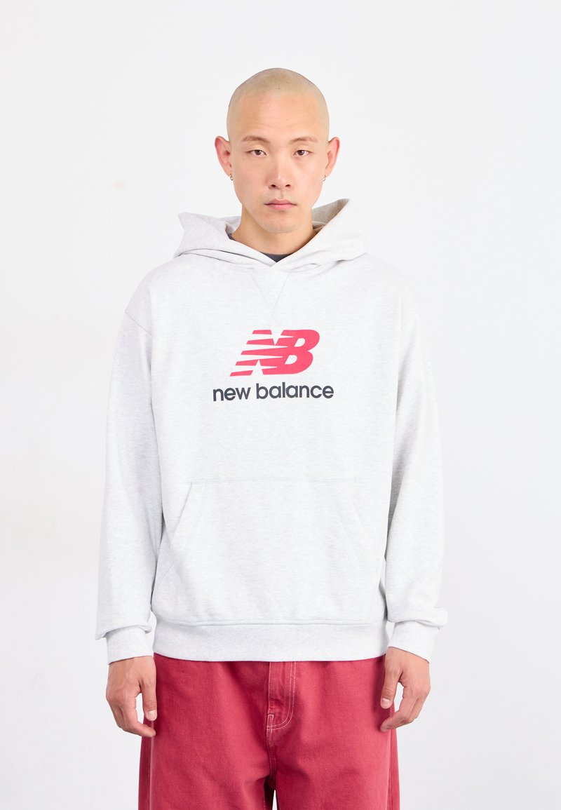 Junger Mann mit rasierter Kopf trägt einen hellgrauen New Balance Kapuzenpullover und rote Hosen, steht vor einem schlichten weißen Hintergrund.
