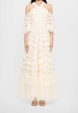 Nainen, joka kantaa kerroksellista, olkapäät paljastavaa maksi-mekkoa, jossa on kerroksia, läpikuultavaa kangasta ja teksturoitu pilkkukuvio, yhdistettynä nudensävyisiin sandaaleihin.