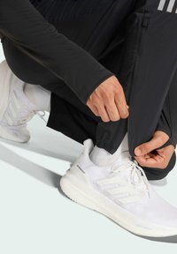 Sorte sportbukser med lynlåse ved anklerne, parret med hvide sneakers med tre striber og tekstureret mesh-overdel. Hænder justerer buksebenet.