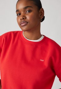 Red sweatshirt made of cotton blend, featuring a crew neck, short sleeves, and a small Levi's logo patch on the chest. Smooth texture.