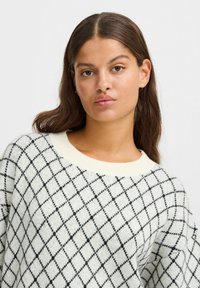 Strickpullover in Creme mit einem schwarzen Diamantenmuster, Rundhalsausschnitt und strukturiertem Finish. Weicher Stoff mit lockerer Passform.