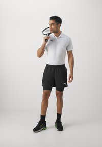 Polo-shirt in het wit, zwarte sportshorts, zwarte sneakers met groene accenten, een tennisracket vasthoudend, staande op een effen achtergrond.