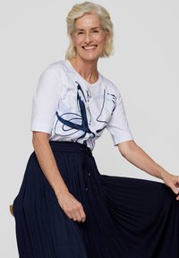 Weißes Baumwoll-T-Shirt mit blauem abstraktem Druck, kombiniert mit einem langen, marineblauen, gefalteten Rock. Das T-Shirt hat kurze Ärmel und einen runden Halsausschnitt.