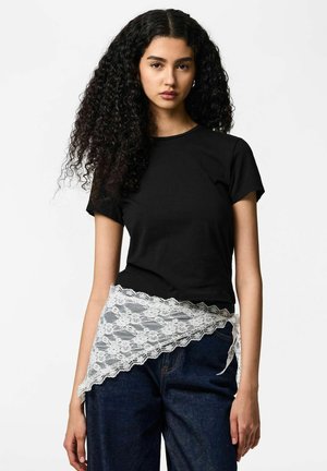 Jeune femme aux cheveux longs et bouclés portant un t-shirt noir, un jean foncé et un cache-cœur en dentelle blanche noué à la taille.