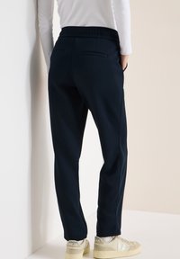 Personne portant un pantalon noir fuselé avec taille élastique et des baskets beiges, debout contre un mur blanc et beige.