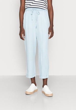 Personne portant un pantalon court bleu clair à cordon, des baskets blanches à plateforme, et un haut rayé noir et blanc, debout devant un fond uni.
