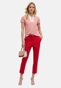 Camisa polo a rayas rojas con cuello blanco, combinada con pantalones rojos ajustados. El modelo sostiene un bolso crema con detalles rojos. Sandalias blancas con tiras.