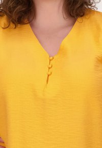 Gele blouse met een V-hals, voorzien van een stolpplooi met drie ronde, met stof overtrokken knopen. Geweven, lichtgewicht materiaal.