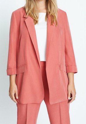 Mujer con una chaqueta blazer color coral con las mangas remangadas, pantalones a juego color coral y una camiseta blanca sobre un fondo blanco.