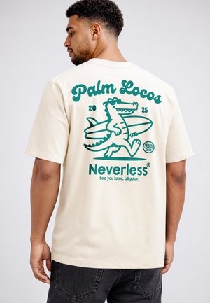 Mann trägt beige T-Shirt mit türkisfarbiger Grafik eines sonnenbrillentragenden Alligators, der ein Surfbrett trägt, und dem Text "Palm Locos 2025 Neverless."